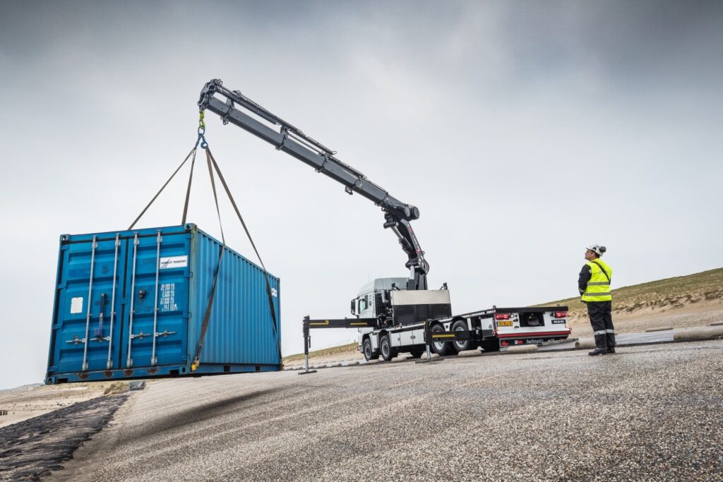 HIAB X-HiPro 1058 loader crane lifting a shipping container from a truck on a coastal site