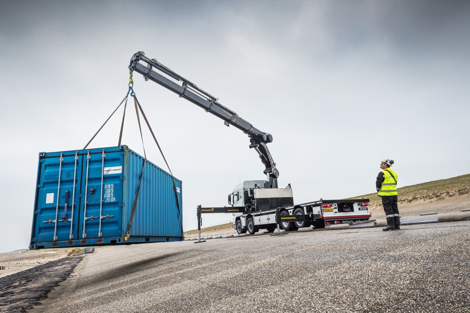 HIAB X-HiPro 1058 loader crane lifting a shipping container from a truck on a coastal site